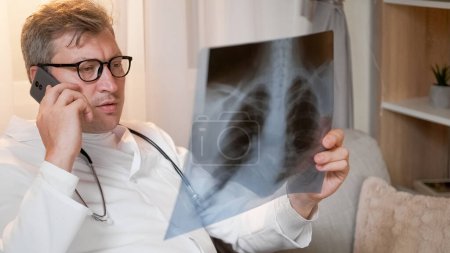 Téléchargez les photos : Consultation mobile. Téléphonez au médecin. Homme spécialiste sérieux avec des poumons film de rayons X parlant de diagnostic sur smartphone assis à l'intérieur de la clinique. - en image libre de droit