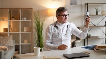 Photo for Medical video advice. Health consultation. Online meeting. Healthcare professional man talking at virtual conference on smartphone sitting at desk in clinic interior. - Royalty Free Image