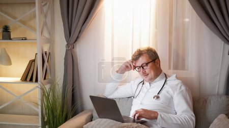 Téléchargez les photos : De nuit. La technologie médicale. Divertissement. Homme détendu dans l'uniforme de l'hôpital défilement ordinateur portable assis sur un canapé confortable à l'intérieur confortable à la fenêtre du soir. - en image libre de droit