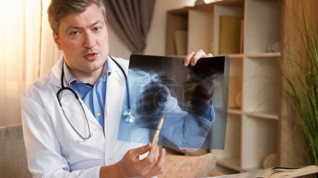 Photo for Diagnosis consultation. Online doctor. Medical specialist man with stethoscope explaining x ray of lungs disease at video appointment sitting at workplace. - Royalty Free Image