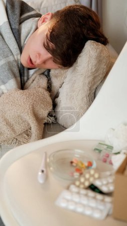 Photo for Seasonal infection. Drugs treatment. Domestic recovery. Sleeping man feeling bad laying in bed covered with plaid with tablets on chair at home. - Royalty Free Image