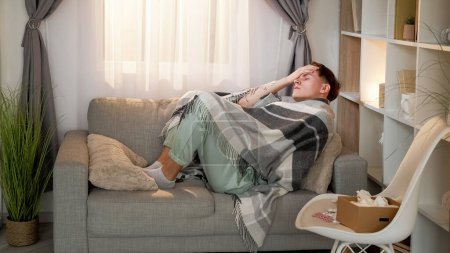 Photo for Sickness fever. Seasonal flu. Ill tired man wrapped in blanket laying on couch suffering from bad headache in living room with drugs at home. - Royalty Free Image