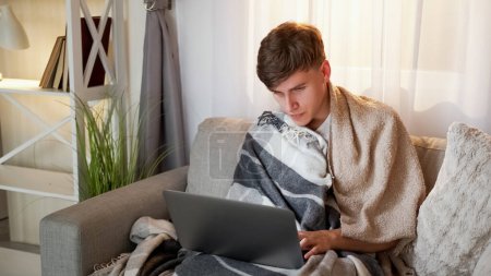 Photo for Distance work. Ill freelancer. Online communication. Fever man on sick leave wrapped in plaid sitting on sofa typing on laptop in living room at home. - Royalty Free Image