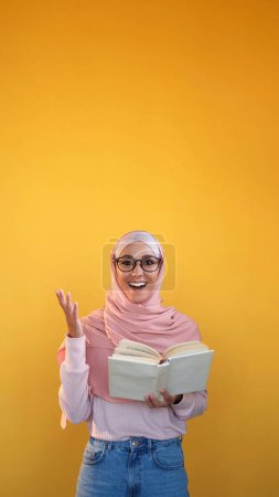Foto de Novela sorprendida. Una lectura interesante. Mujer asombrada en hijab sosteniendo libro increíble texto de la trama aislado en el fondo del espacio de copia amarillo. - Imagen libre de derechos