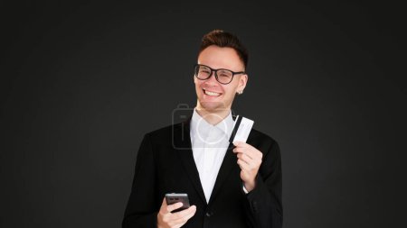 Photo for Mobile banking. Online e-commerce. Cheerful satisfied business man holding phone credit card isolated on dark black empty space background. - Royalty Free Image