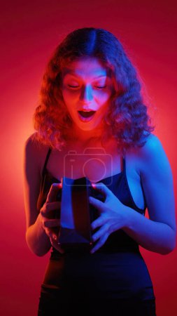 Photo for Present surprise. Special gift. Shocked delighted young woman with glowing face opening paper package isolated on pink spotlight background. - Royalty Free Image