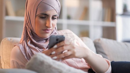 Spam message. Internet bullying. Frustrated grumpy woman in hijab texting on phone upset with communication problem bad news on sofa at home.