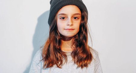 Child fashion. Urban kid. Confident relaxed cute little girl model in black knitted beanie hat on white shadow light free space background.