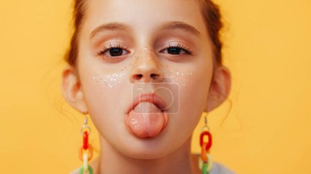 Photo for Tongue out child. Grimacing kid. Naughty playful little girl with fake glitter freckles on cute face mocking isolated on yellow background. - Royalty Free Image
