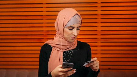Foto de Pago móvil. Compras por Internet. Mujer atractiva en hijab ordenar compra en línea en la aplicación de banca telefónica con transferencia de dinero de la tarjeta de crédito en la cafetería. - Imagen libre de derechos