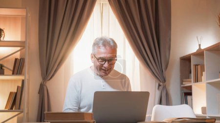 Online communication. Mature man. Digital life. Happy senior male working laptop on desk light home interior.