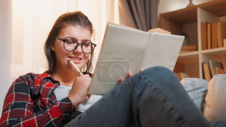 Téléchargez les photos : Étudiant. Joie aux devoirs. Souriante femme appréciant l'examen de livre apprendre heureux avec la leçon de lycée sur le canapé à la maison. - en image libre de droit