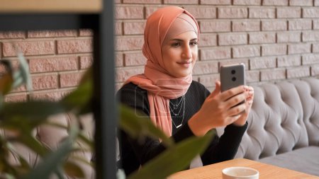 Photo for Mobile using. Video call. Happy woman in hijab speaking to friend on cellphone internet conversation checking social network at cozy coffee shop. - Royalty Free Image