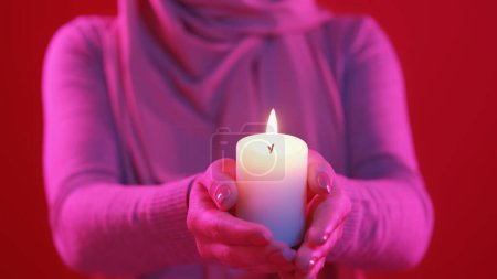 Téléchargez les photos : Un feu de prière. Vacances islamiques. Lumière de bougie. Femme méconnaissable dans le hijab tenant flamme de cire brûlante dans la lueur rose vif néon sur fond rouge. - en image libre de droit