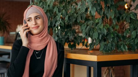 Conversación móvil. Conversación telefónica. Mujer feliz en hijab hablando en el teléfono inteligente agradable llamada amistosa en el acogedor café espacio libre.