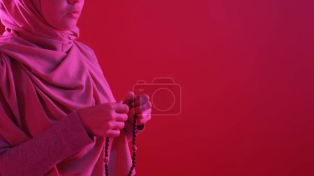 Photo for Islamic praying. Traditional tasbih. Unrecognizable woman with wooden rosary beads in vibrant pink neon light on red empty space background. - Royalty Free Image