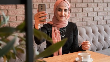 Photo for Cafe selfie. Online stream. Pretty woman in hijab making photo creating content for blog internet conversation at cozy coffee shop. - Royalty Free Image