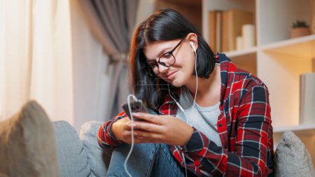 Photo for Phone chatting. Gadget leisure. Happy millennial woman typing message on smartphone listening song in earphones home light interior. - Royalty Free Image
