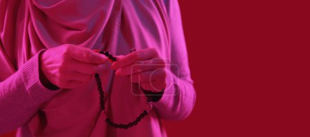 Téléchargez les photos : Beads praying. Tasbih tradition.Closeup of woman hands with wooden rosary islamic belief pink neon light on red copy space background. - en image libre de droit