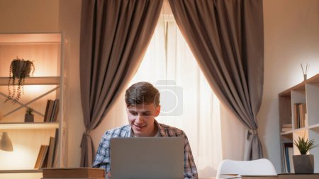 Online education. Male student. Digital research. Happy man with books on desk looking laptop light home interior.