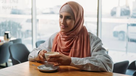 Cafe relax. Coffee break. Serene dreamy woman in hijab drinking cup of hot beverage at window enjoying leisure in cozy coffeehouse.