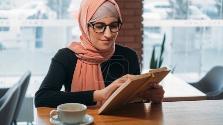 Foto de Libro del café. Leyendo ocio. Mujer concentrada en hijab y gafas estudiante universitaria estudiando novela de aprendizaje en acogedora cafetería espacio libre. - Imagen libre de derechos