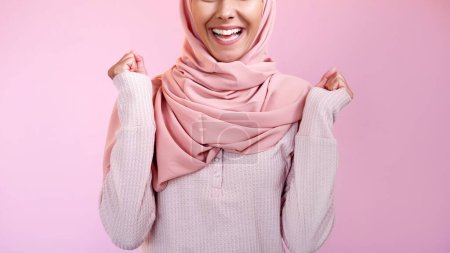 Winner yes. Victory joy. Positive reaction. Unrecognizable pleased satisfied excited smiling happy woman in hijab isolated on pink copy space background.