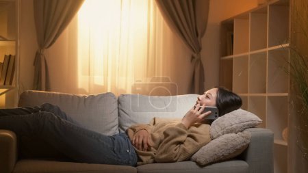 Téléchargez les photos : Phone talk. Home leisure. Weekend communication. Relaxed woman speaking lying on cozy couch in living room interior. - en image libre de droit