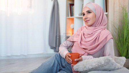 Foto de Contemplación domiciliaria. Relajación fin de semana. Mujer pensativa inspirada en hijab con taza de café disfrutando de bebida caliente soñando solo en el interior de la sala de estar moderna con espacio libre. - Imagen libre de derechos
