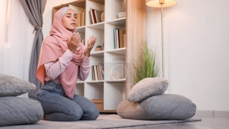 Téléchargez les photos : Je prie musulman. Islam religion. Allah adore. Femme fidèle en hijab lisant le Saint Coran sur le sol à la lumière intérieur de la maison moderne avec espace libre. - en image libre de droit