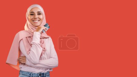 Bank service. Electronic money. Cheerful pleased woman in headscarf holding credit card isolated on red empty space advertising background.