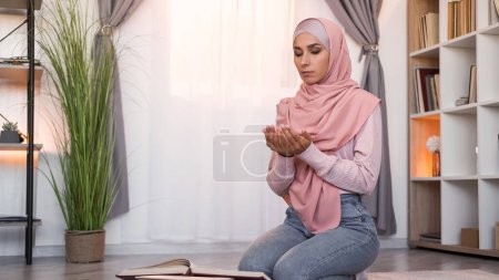 Photo for Koran praying. Faithful muslim. Islam culture. Calm focused woman in hijab with closed eyes reading Holy Quran at Ramadan on floor at light modern home interior with free space. - Royalty Free Image