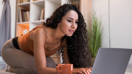 Online-Chat. Virtuelle Freizeit. Internetkommunikation. Neugierige Frau tippt mit Laptop auf dem Boden zu Hause Wohnzimmer Innenraum mit freiem Platz.