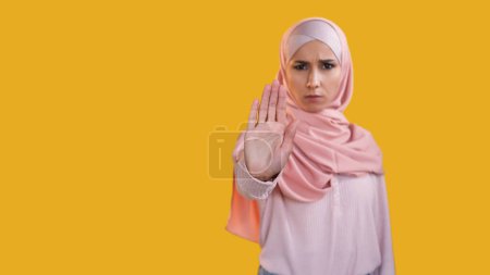 Foto de Alto la mano. Sin gestos. Mujer seria asustada perturbada en alerta de hijab con el cartel de protesta de la palma extendida aislada en el fondo amarillo desenfocado del espacio vacío. - Imagen libre de derechos