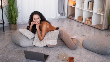 Loisirs Internet. Week-end en ligne. Détendu curieuse fille souriante profiter de regarder la vidéo sur ordinateur portable sur le sol à l'intérieur de la maison moderne avec espace libre.