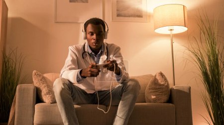 Photo for Gaming addiction. Virtual leisure. Digital lifestyle. Disturbed male doctor in headphones playing on phone on night shift on couch at modern evening home interior with free space. - Royalty Free Image