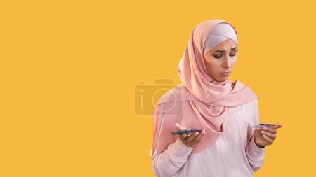 Payment error. Bank fraud. Transaction fail. Confused puzzled woman in hijab using mobile phone credit card isolated on orange empty space background.