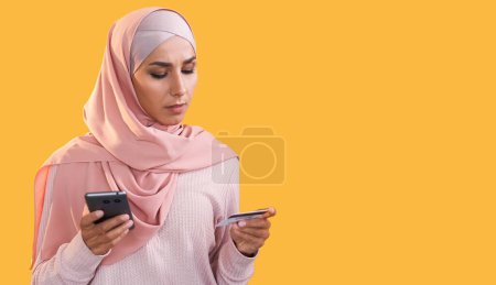 Foto de Mobile banking. Online shopping. Serious woman in hijab using credit card phone for digital money transaction isolated on orange empty space background. - Imagen libre de derechos