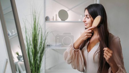 Hilos de belleza. Cuidado matutino. Un tratamiento saludable. Mujer bastante elegante cepillado pelo largo morena en el espejo en el hogar luz espacio de copia interior.