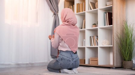 Téléchargez les photos : La prière islamique. La foi musulmane. Vue arrière d'une femme religieuse méconnaissable dans le hijab lisant la louange du Saint Coran à Allah sur le sol à la lumière intérieur de la maison moderne avec espace libre. - en image libre de droit