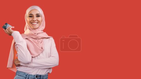 Pago sin efectivo. Servicio bancario. Transferencia electrónica. Feliz mujer sonriente satisfecha en hijab con tarjeta de crédito aislada sobre fondo rojo vacío.