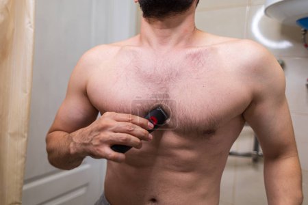 Foto de Un hombre quita el pelo no deseado de su cuerpo usando una afeitadora eléctrica. Un hombre con una figura atlética en el baño. - Imagen libre de derechos