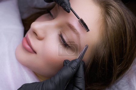 Photo for The master uses tweezers to correct the shape of the eyebrows before the permanent makeup procedure. PMU Procedure, Permanent Eyebrow Makeup. - Royalty Free Image