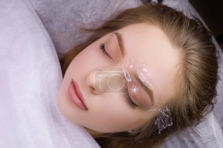 Photo for The model's eyebrows are anesthetized and one eyebrow is covered with a clear protective polyethylene. PMU Procedure, Permanent Eyebrow Makeup. - Royalty Free Image