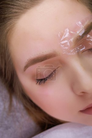 The model's eyebrows are anesthetized and one eyebrow is covered with a clear protective polyethylene. PMU Procedure, Permanent Eyebrow Makeup.