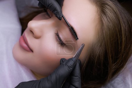 Plucking the eyebrows along the contour with tweezers before the permanent makeup procedure. PMU Procedure, Permanent Eyebrow Makeup.