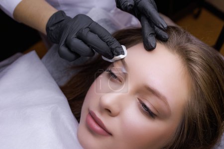 Photo for A close-up of the master wiping the model's eyebrows before the permanent makeup procedure. PMU Procedure, Permanent Eyebrow Makeup. - Royalty Free Image