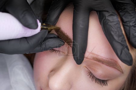 Nahaufnahme des tätowierenden Pigments in die Haut der Augenbrauen mit einem Tätowiergerät. PMU-Verfahren, Permanentes Augenbrauen-Make-up.