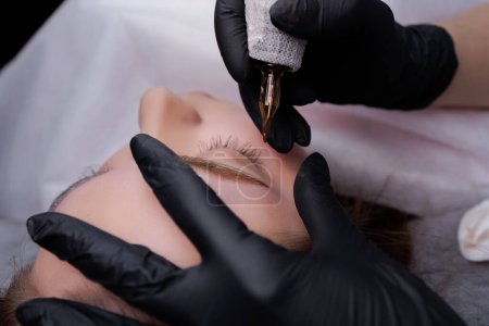 Photo for In the hands of the tattoo master is a machine in which a cartridge is inserted that applies pigment to the skin of the eyebrows. PMU Procedure, Permanent Eyebrow Makeup. - Royalty Free Image
