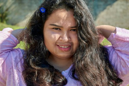 Portrait of a young Latin American woman on a summer day. She has long black hair. She is smiling at the camera. She has a blue flower in her hair.